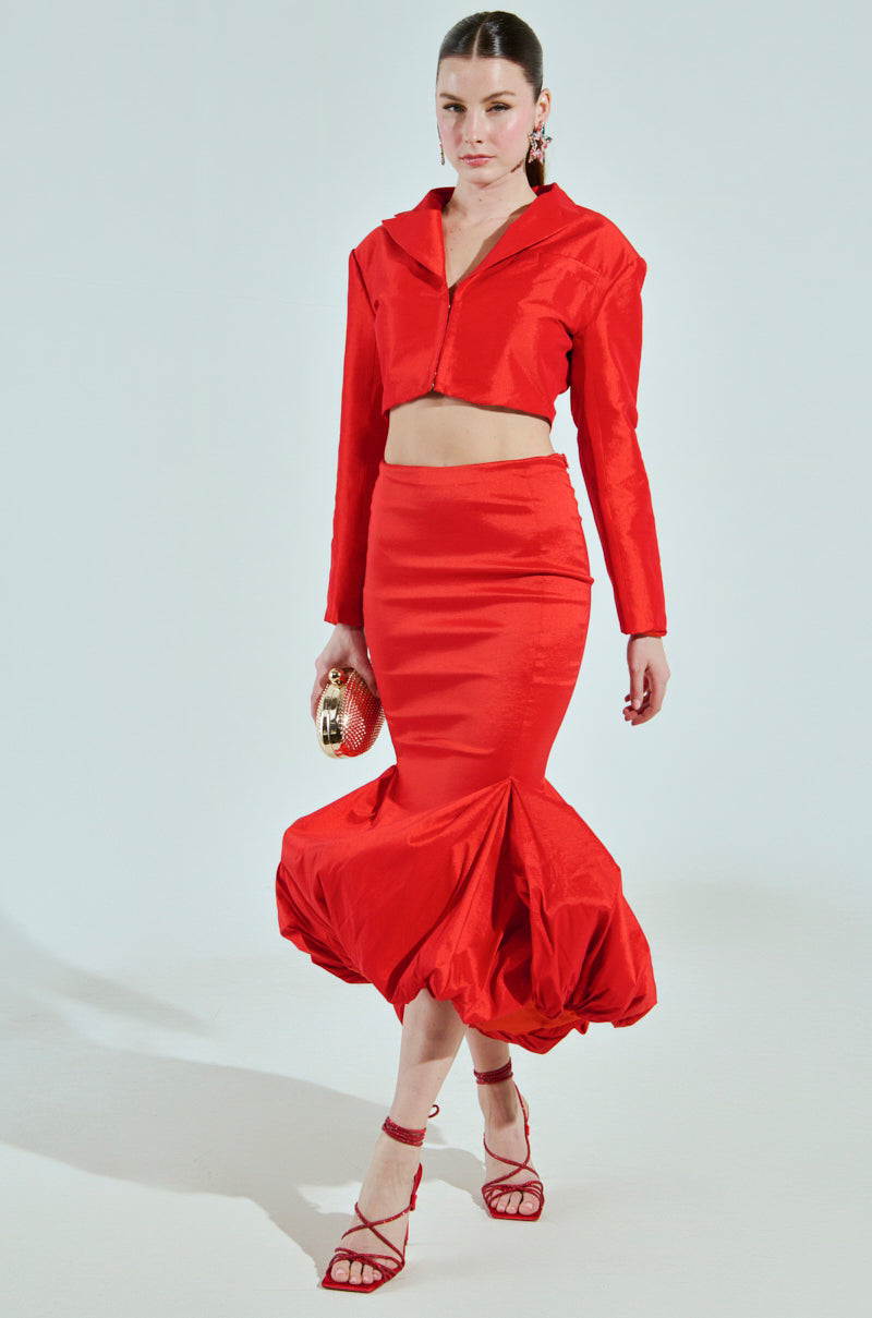 A model confidently showcases a cropped boxy silhouette in a vibrant red outfit, wearing a fitted skirt with a flared hem. She accents her look with red strappy heels and a small clutch, her hair neatly pulled back, spotlighting the ensemble's elegance like the BRIGHTEST LIGHT BLAZER.