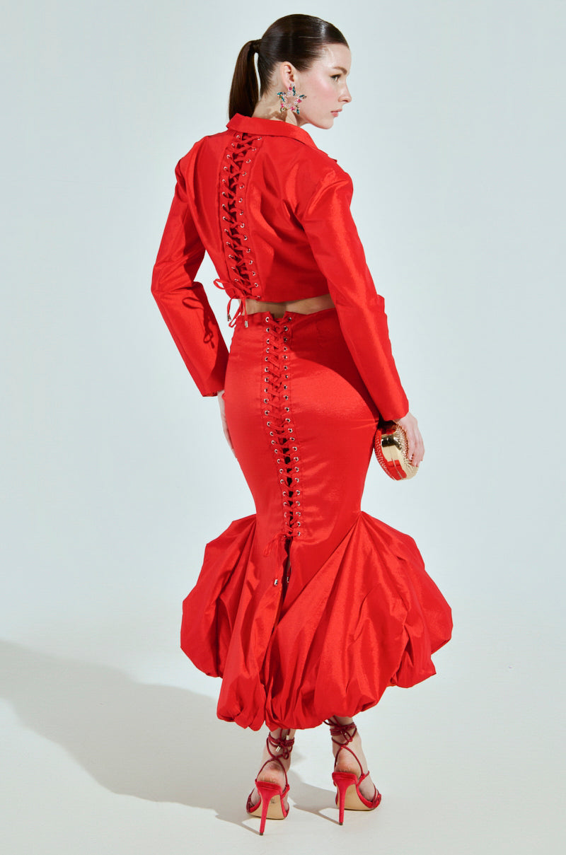 A woman in profile showcases the BRIGHTEST LIGHT BLAZER in red, featuring lace-up details on the back and skirt. The cropped boxy silhouette pairs with a flared skirt and red high heels. She holds a small gold clutch, her hair elegantly tied back.
