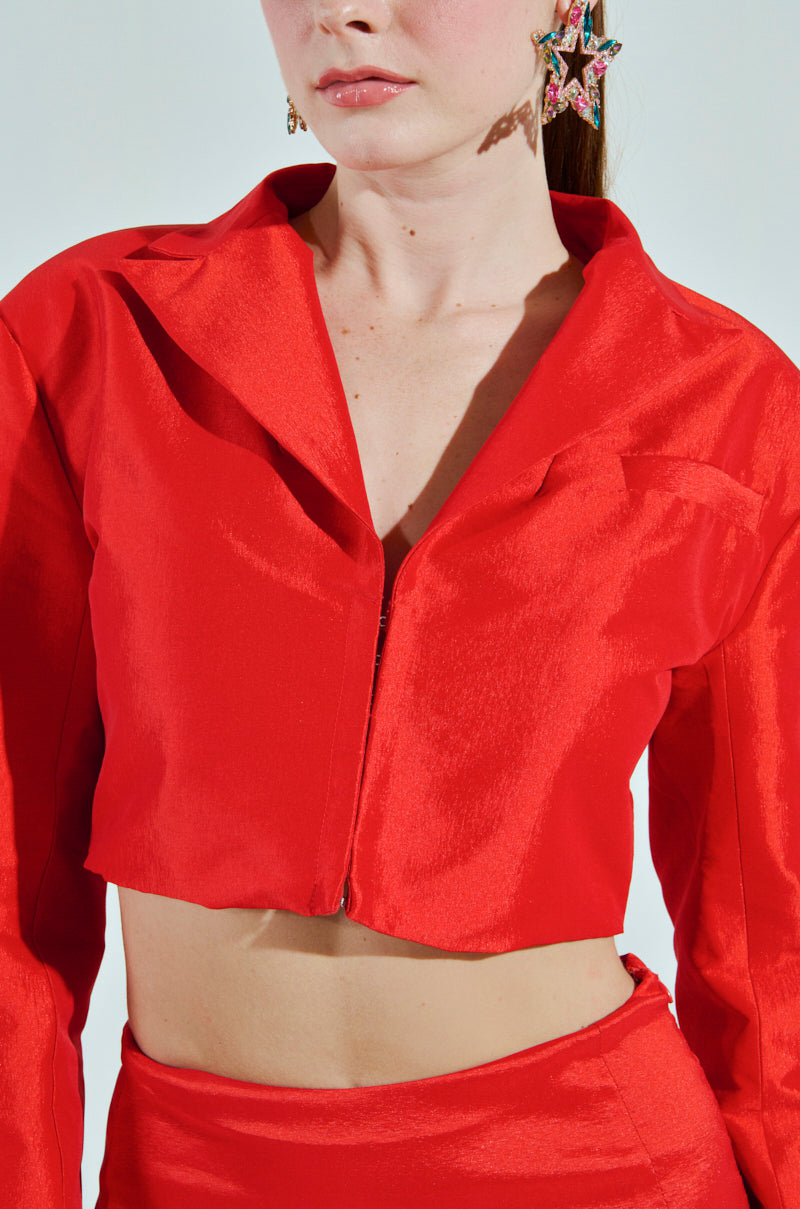 A woman dressed in the BRIGHTEST LIGHT BLAZER—a bright red satin outfit with a cropped boxy jacket and high-waisted pants—wears star-shaped earrings. She stands against a white background, her face partially visible, showing a neutral expression.