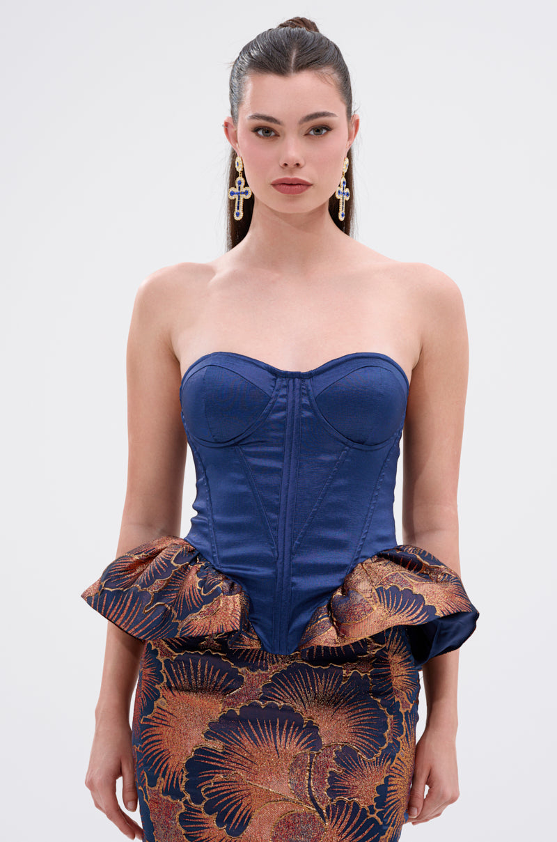 A woman with long, dark hair wears the LADY LUX PEPLUM BROCADE CORSET TOP in bronze and blue, styled with a matching floral skirt and bold earrings, standing against a simple light background.
