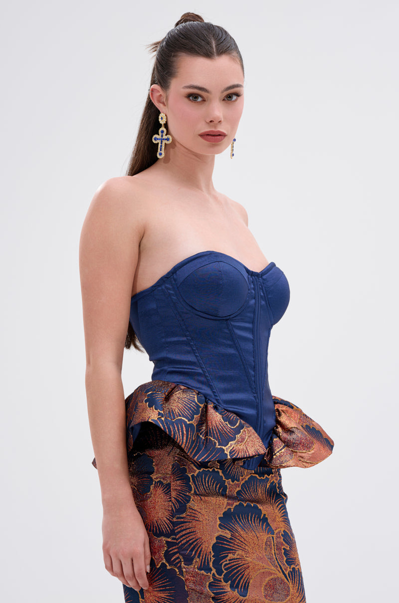 A woman with long brown hair in a high ponytail wears the LADY LUX PEPLUM BROCADE CORSET TOP, a strapless blue corset with structured seams and peplum detail, paired with a patterned blue, rust, and gold skirt and cross-shaped earrings.