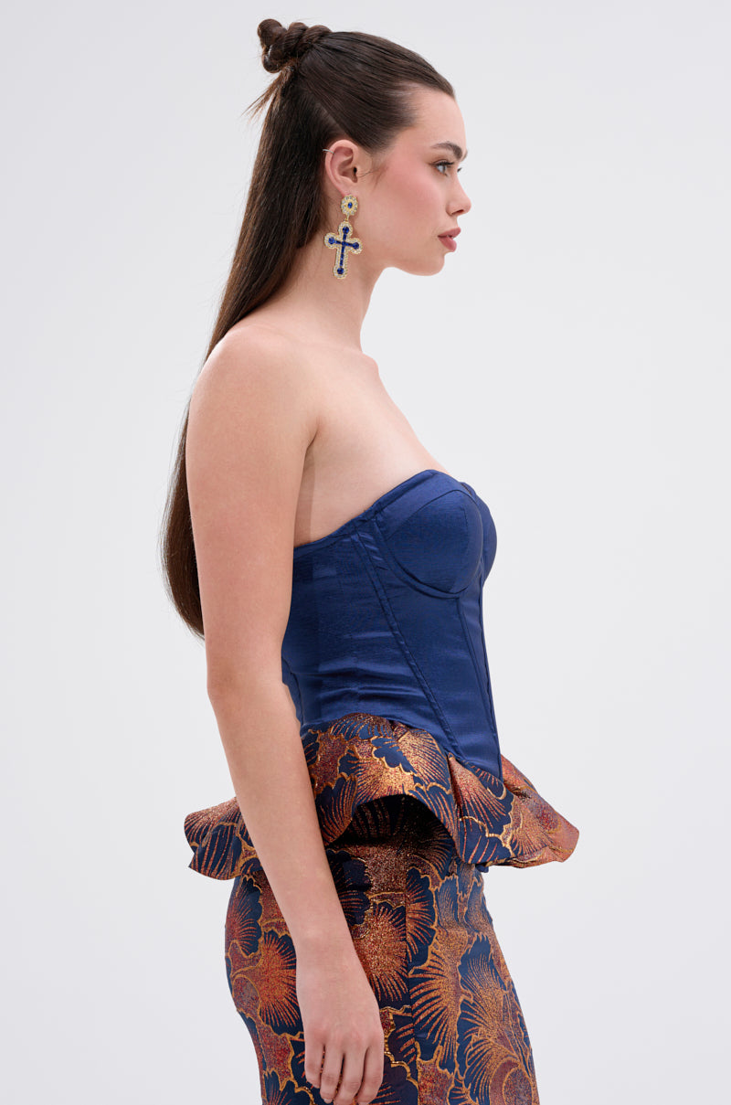 A woman with long, straight brown hair wears the LADY LUX PEPLUM BROCADE CORSET TOP in royal blue, featuring a strapless silhouette and flared peplum brocade hem, paired with a matching skirt and bold cross earrings against a plain background.
