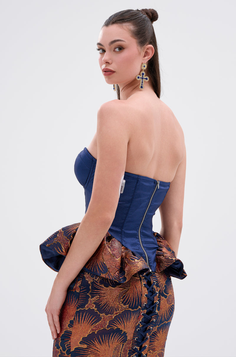 A woman with dark hair in a high ponytail models the LADY LUX PEPLUM BROCADE CORSET TOP, a strapless blue design with gold and orange floral patterns, peplum ruffles, and brocade detail. She stands sideways, looking over her shoulder at the camera.