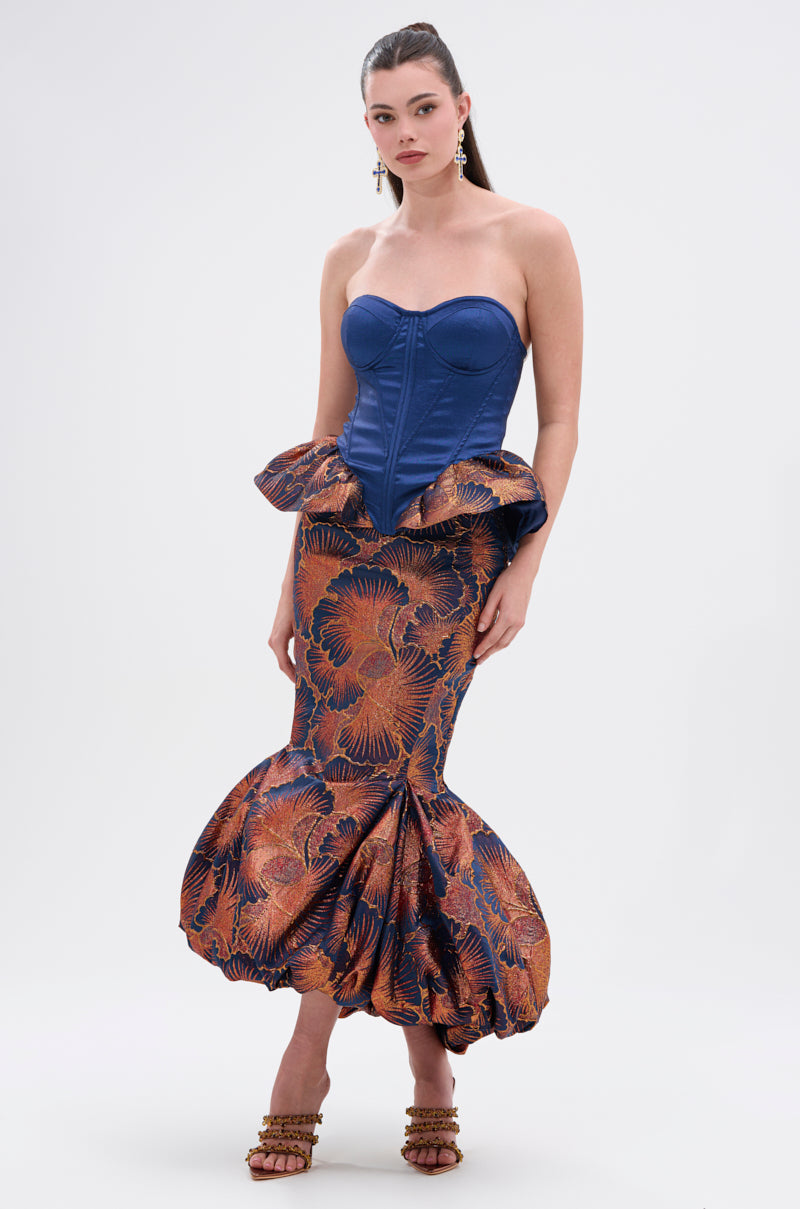 A woman models the LADY LUX PEPLUM BROCADE CORSET TOP, a strapless silhouette with peplum detail, paired with a mermaid skirt in bold orange and blue floral print. She accessorizes with dangling earrings, brown heels, and slicked-back hair.