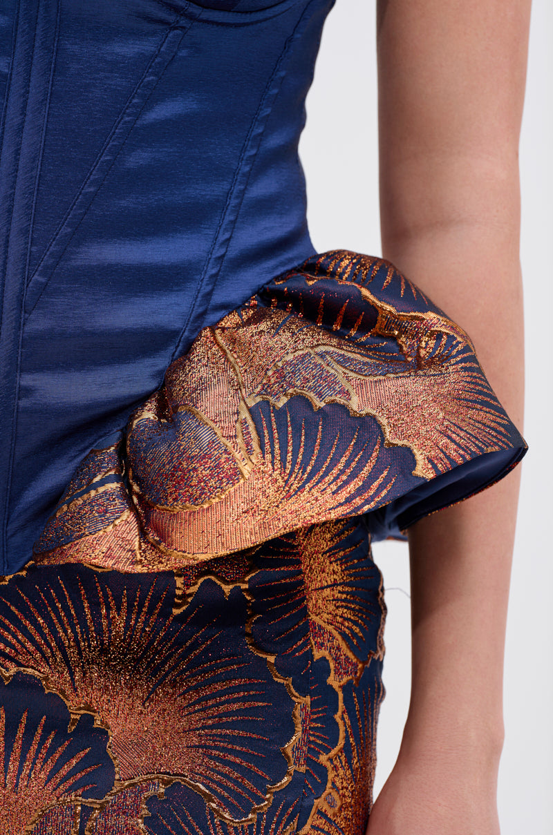 Close-up of a person wearing the LADY LUX PEPLUM BROCADE CORSET TOP, featuring a shiny blue strapless design paired with a textured peplum brocade skirt, adorned with metallic gold and navy floral patterns and ruffled waist detailing.