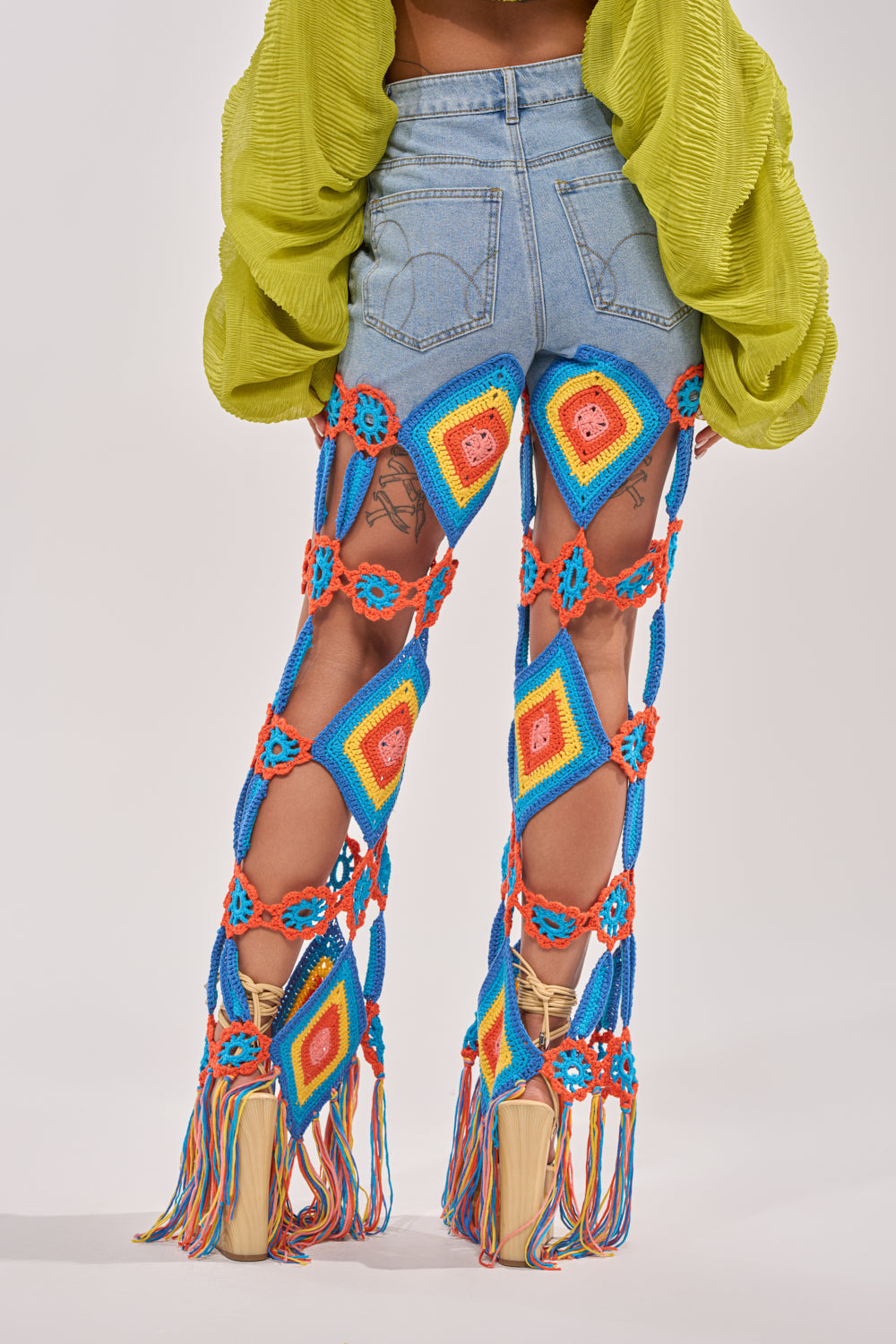 Someone faces away, wearing OUT OF OFFICE CROCHET DENIM JEANS with vibrant crochet squares and tassels, styled with a bright green textured top and beige platform shoes for a standout festival look. 3