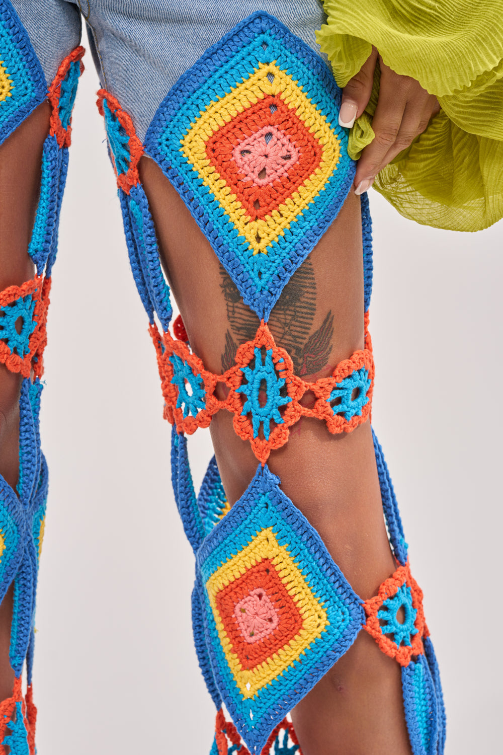 Close-up of a person wearing OUT OF OFFICE CROCHET DENIM JEANS featuring vibrant blue, yellow, orange, and red crochet squares that reveal parts of the thigh and a feather tattoo—perfect for making a bold festival statement. 8