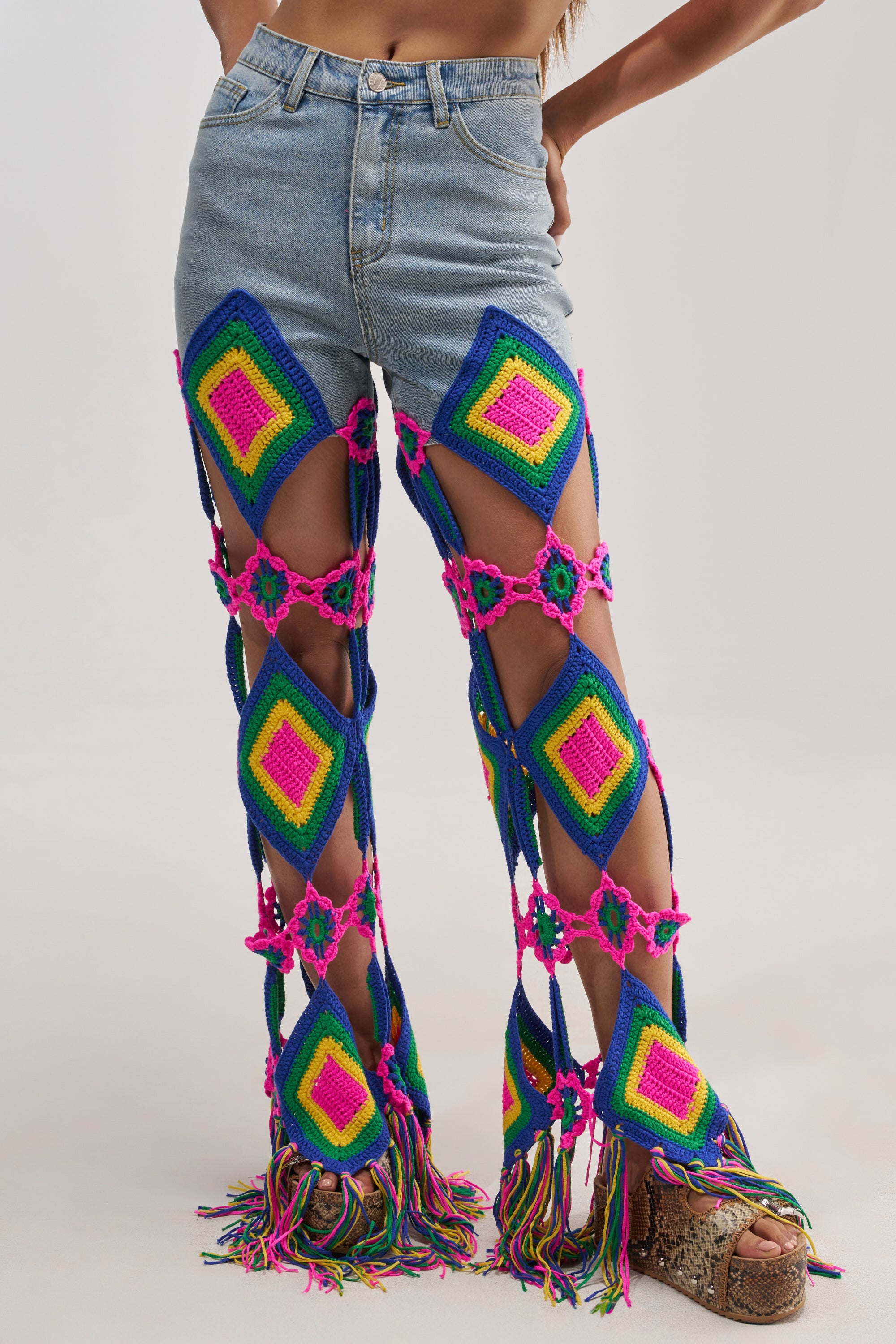 A person wearing OUT OF OFFICE CROCHET DENIM JEANS, featuring high-waisted denim with colorful crocheted panels, large geometric cutouts that reveal skin, and fringe extending down the legs for a festival-inspired look.