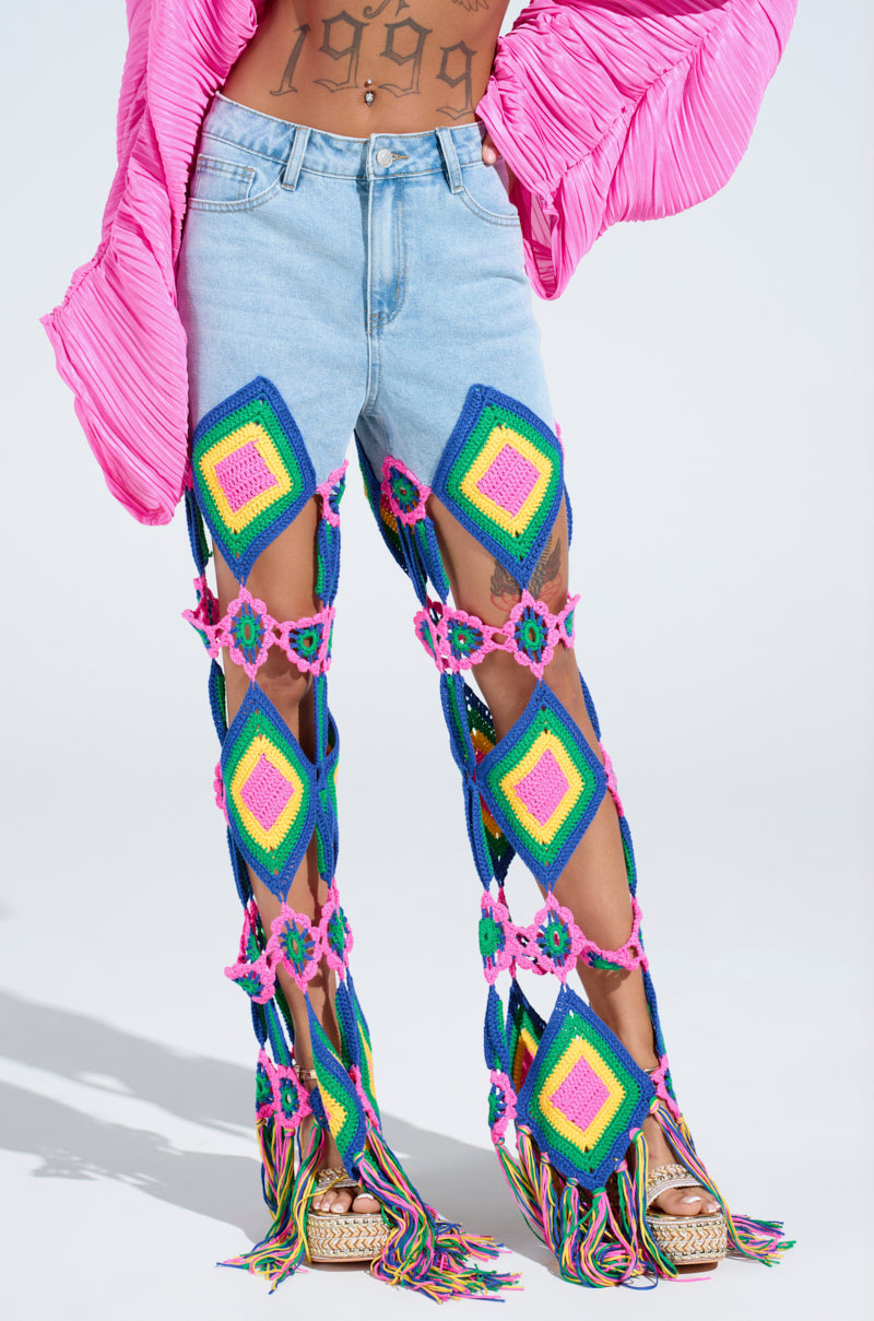 Wearing OUT OF OFFICE CROCHET DENIM JEANS, styled with granny square motifs, a person sports a pink textured top and tattooed midriff, completing the look with platform sandals featuring rainbow fringe hems.