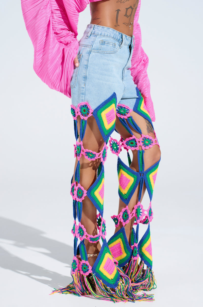 Wearing light blue denim shorts, accented by colorful crochet leg coverings with granny square designs and rainbow fringe hems, the individual pairs them with a bright pink long-sleeved top. The scene is set against a soft, light backdrop.