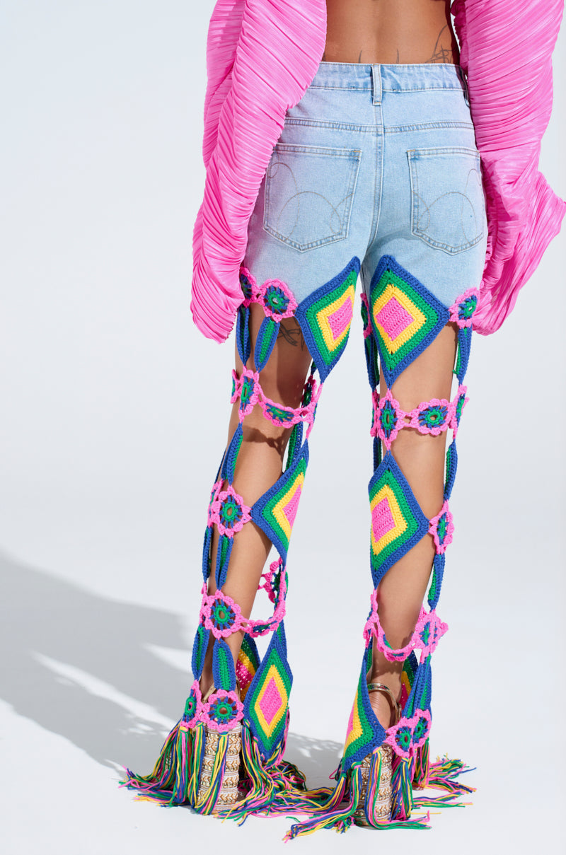 Someone wearing OUT OF OFFICE CROCHET DENIM JEANS with colorful granny square motifs in vibrant pink, green, and yellow diamonds. The jeans have rainbow fringe hems and are styled with a bright pink top against a plain white backdrop.