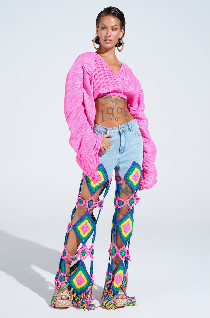Amidst a plain backdrop, an individual exudes confidence in a vibrant ensemble with a pink textured top and OUT OF OFFICE CROCHET DENIM JEANS featuring distinctive rainbow fringe hems. Their tattooed arms highlight a bold "1998" on the stomach.