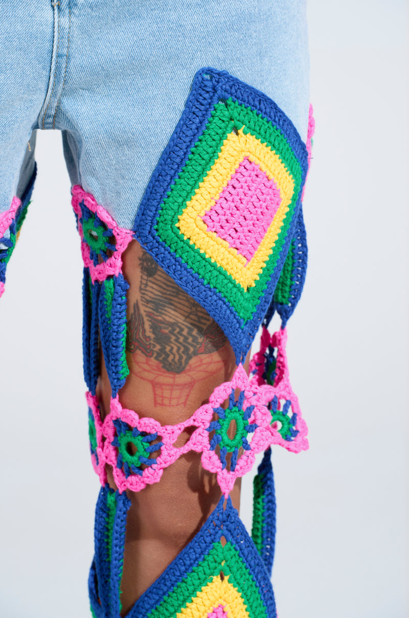 A close-up of a leg wearing OUT OF OFFICE CROCHET DENIM JEANS shows vibrant granny square motifs and flowers on light blue fabric, with a tattoo visible through open spaces, enhancing the eye-catching design.