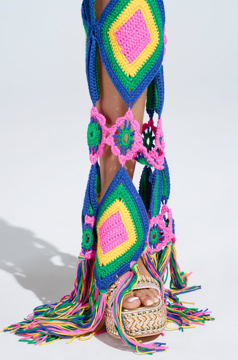 The OUT OF OFFICE CROCHET DENIM JEANS display diamond shapes, floral patterns, and granny square motifs in pink, blue, yellow, and green with rainbow fringe hems. Styled with tan platform sandals against a light backdrop for added flair.