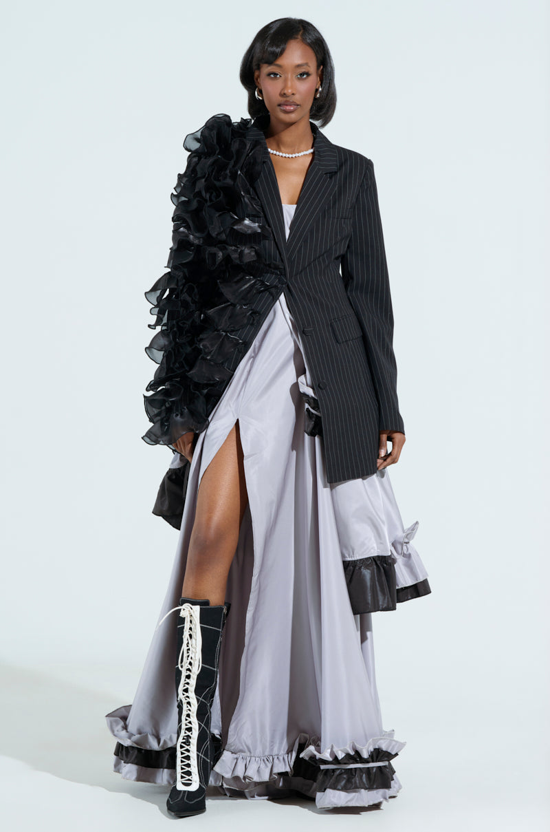A woman showcases the RAMY RUFFLE BLAZER with tonal ruffles and pinstripes, paired with a gray slit dress and black high-heeled boots with white laces. She completes her chic outfit with a pearl necklace.
