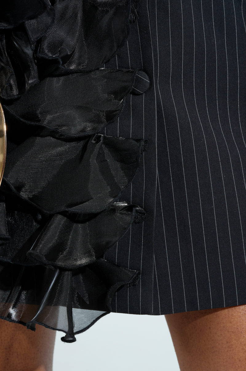 Close-up of the RAMY RUFFLE BLAZER featuring black pinstripes and black satin tonal ruffles on the side, capturing a blend of textures and patterns. The frame shows part of a person wearing it.
