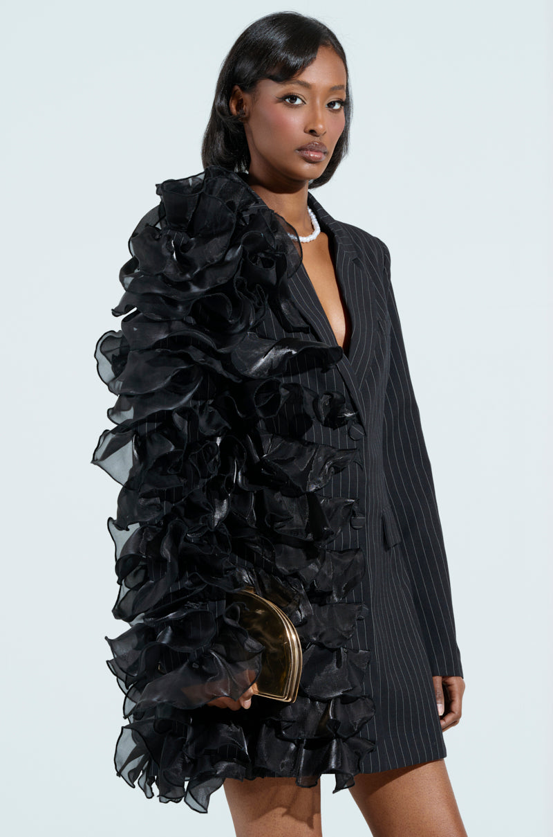 A woman stands against a light background wearing the RAMY RUFFLE BLAZER, with dramatic black ruffles cascading down one side. She holds a small gold clutch and accessorizes her sleek, shoulder-length hair with a pearl necklace.
