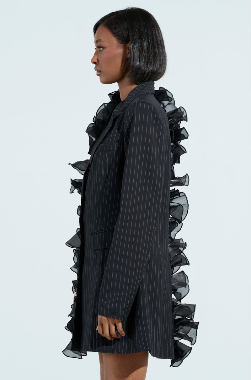 Against a light background, an individual with short hair stands in profile wearing the RAMY RUFFLE BLAZER. The pinstripe design perfectly matches the black ruffles that elegantly enhance both the front and back edges of this sophisticated piece.