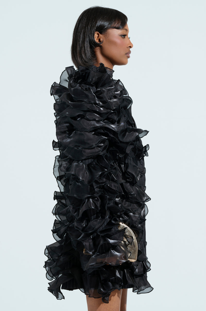 A woman with short dark hair poses in profile wearing the RAMY RUFFLE BLAZER, a textured black blazer with tonal ruffles. The voluminous design complements her silhouette as she stands against a plain light blue background.