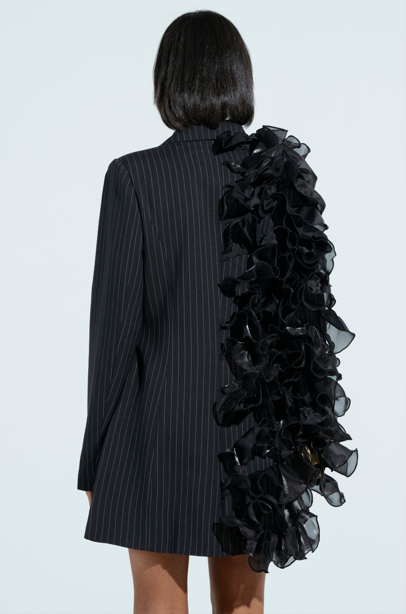 Facing away against a plain light blue background, a person wears the RAMY RUFFLE BLAZER, featuring an asymmetrical design with cascading black ruffles on one side and a pinstripe pattern that complements the piece beautifully.