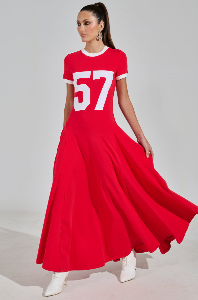 A woman in a COOLEST IN THE ROOM Jersey Maxi Dress, with the number 57, paired with white ankle boots. She stands against a plain backdrop, her hair in a sleek ponytail, confidently looking to the side.