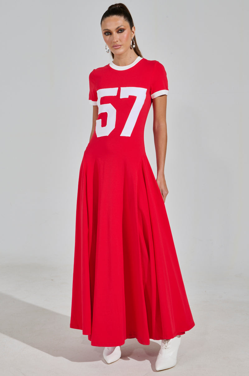 A woman models the COOLEST IN THE ROOM JERSEY MAXI DRESS, a vibrant red maxi featuring a white "57" on the front. With short sleeves and a fitted bodice, she pairs it with chic white ankle boots against a plain backdrop for an ideal game day look.