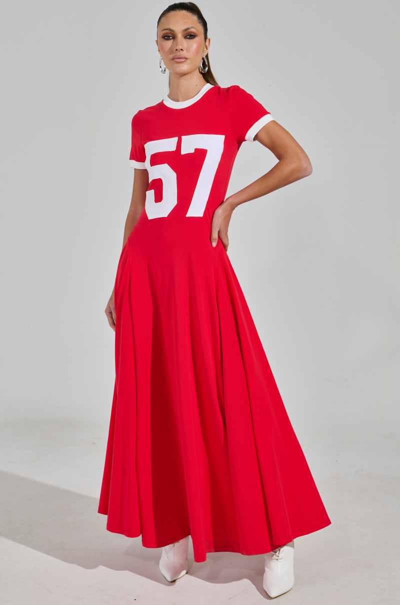 Wearing the COOLEST IN THE ROOM JERSEY MAXI DRESS, featuring a red design with a white "57" on the front and short sleeves, stands against a neutral background. The ensemble is complemented by stylish white shoes, making it perfect for game day.