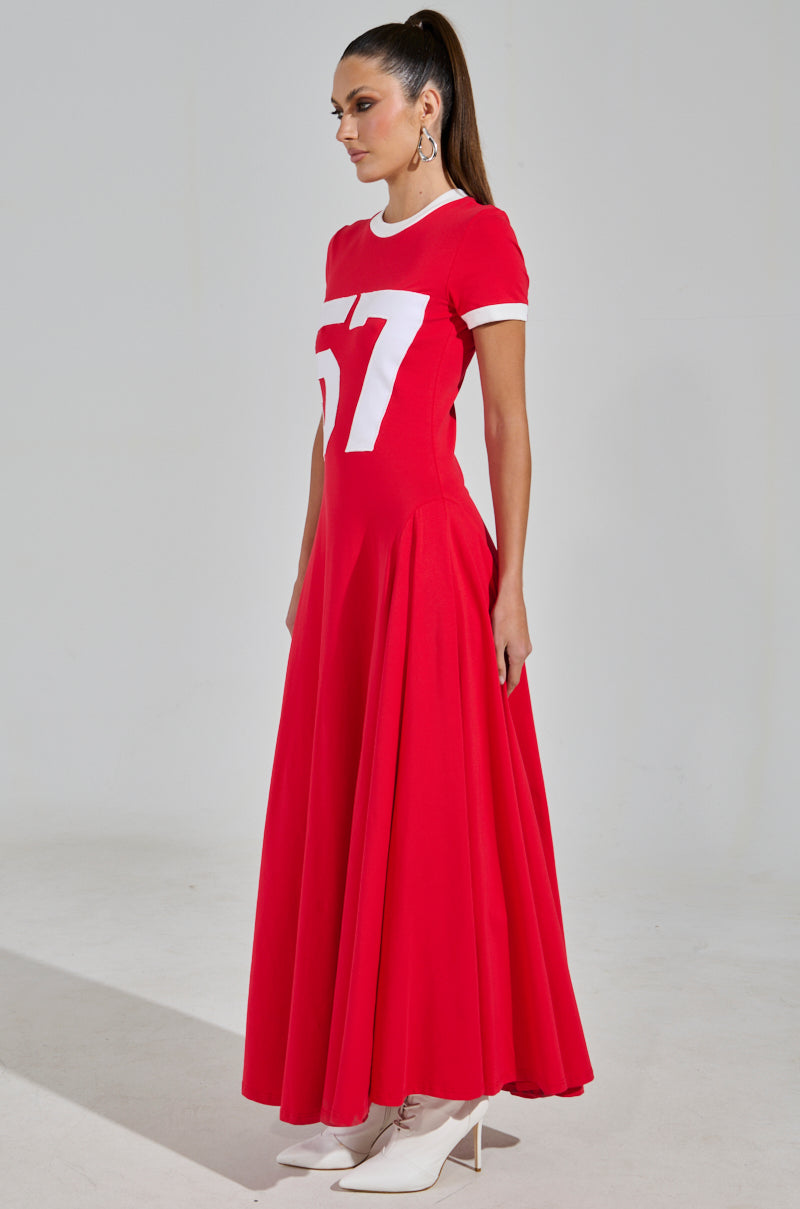 The woman is wearing the COOLEST IN THE ROOM JERSEY MAXI DRESS, a sleek red dress with "57" in white on the front. Her high ponytail adds flair, while white ankle boots complete her game day look against the plain backdrop.