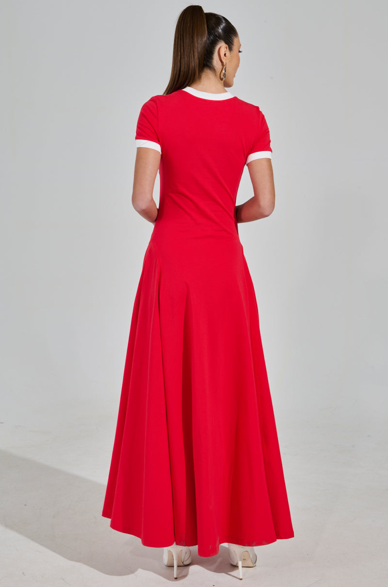 A person with long hair in a ponytail wears the COOLEST IN THE ROOM JERSEY MAXI DRESS, featuring white-trimmed short sleeves and a flowing maxi skirt. They pair this striking red dress with white heels, perfectly embodying an elegant game day ensemble against a plain backdrop.