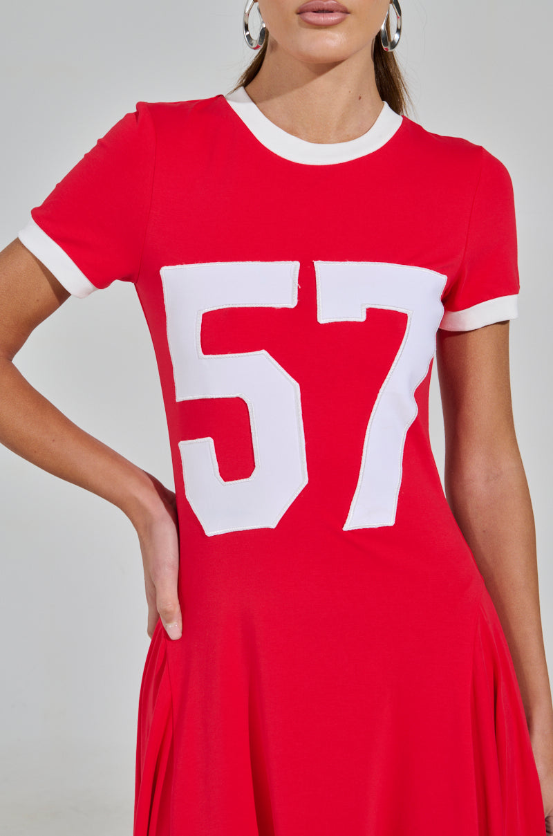 A person confidently poses in the COOLEST IN THE ROOM JERSEY MAXI DRESS, featuring a red design with a white number 57 on the front, standing against a plain light background that highlights this game day ensemble.