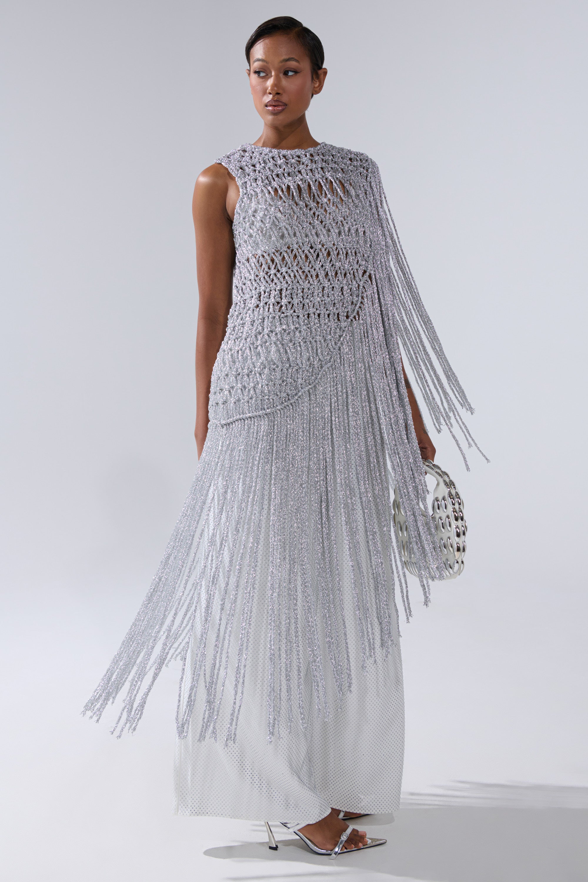 A woman models the UNDRESS FRINGE BLOUSE, a sleeveless silvery knit top with long fringe, styled over a white floor-length dress. She accessorizes with a metallic handbag and pointed silver heels against a plain light gray backdrop.