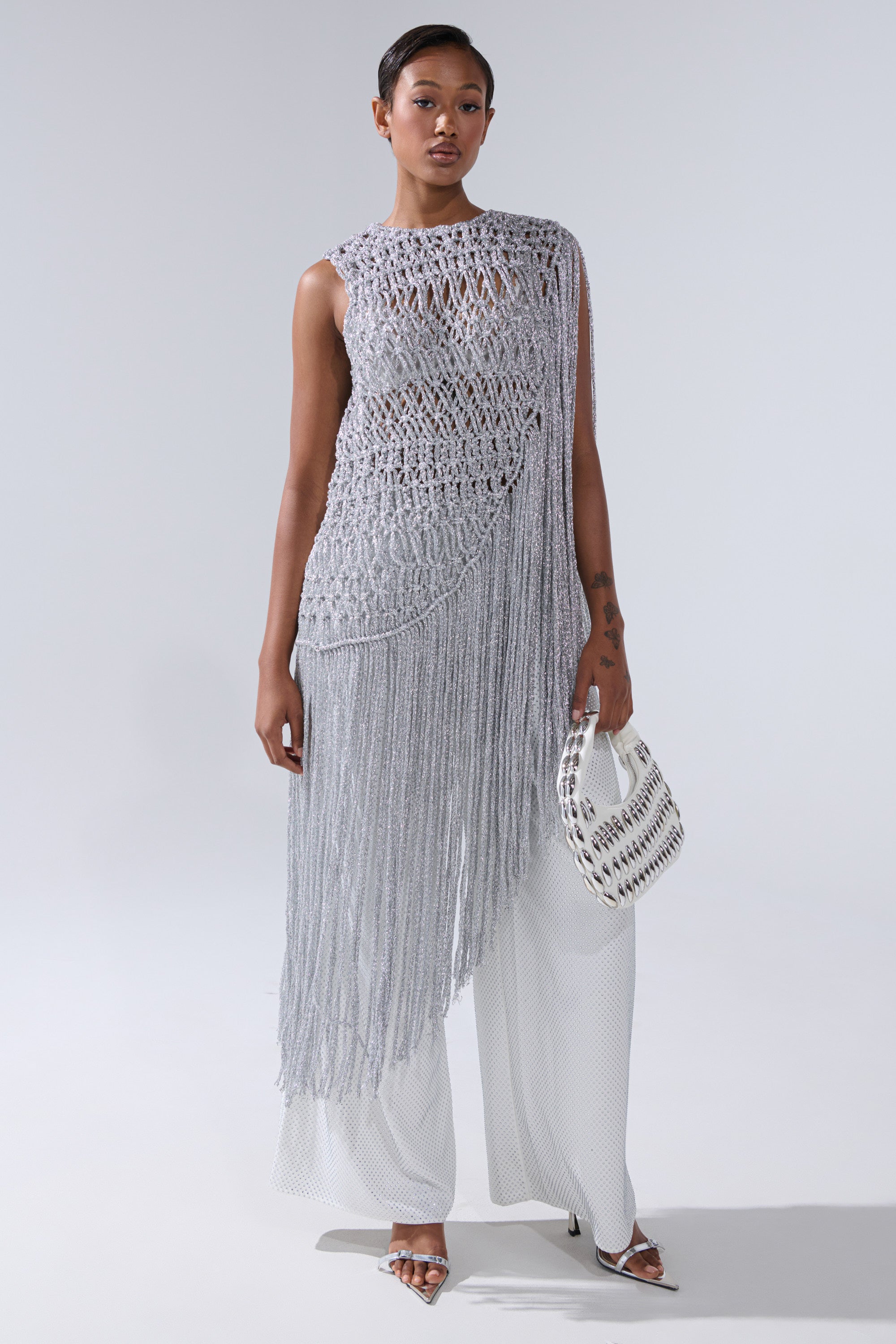 A woman models the UNDRESS FRINGE BLOUSE—a sleeveless, asymmetrical silver crochet top with long fringe—styled over white wide-leg pants, accessorized with a small textured handbag and strappy heels against a plain light background.