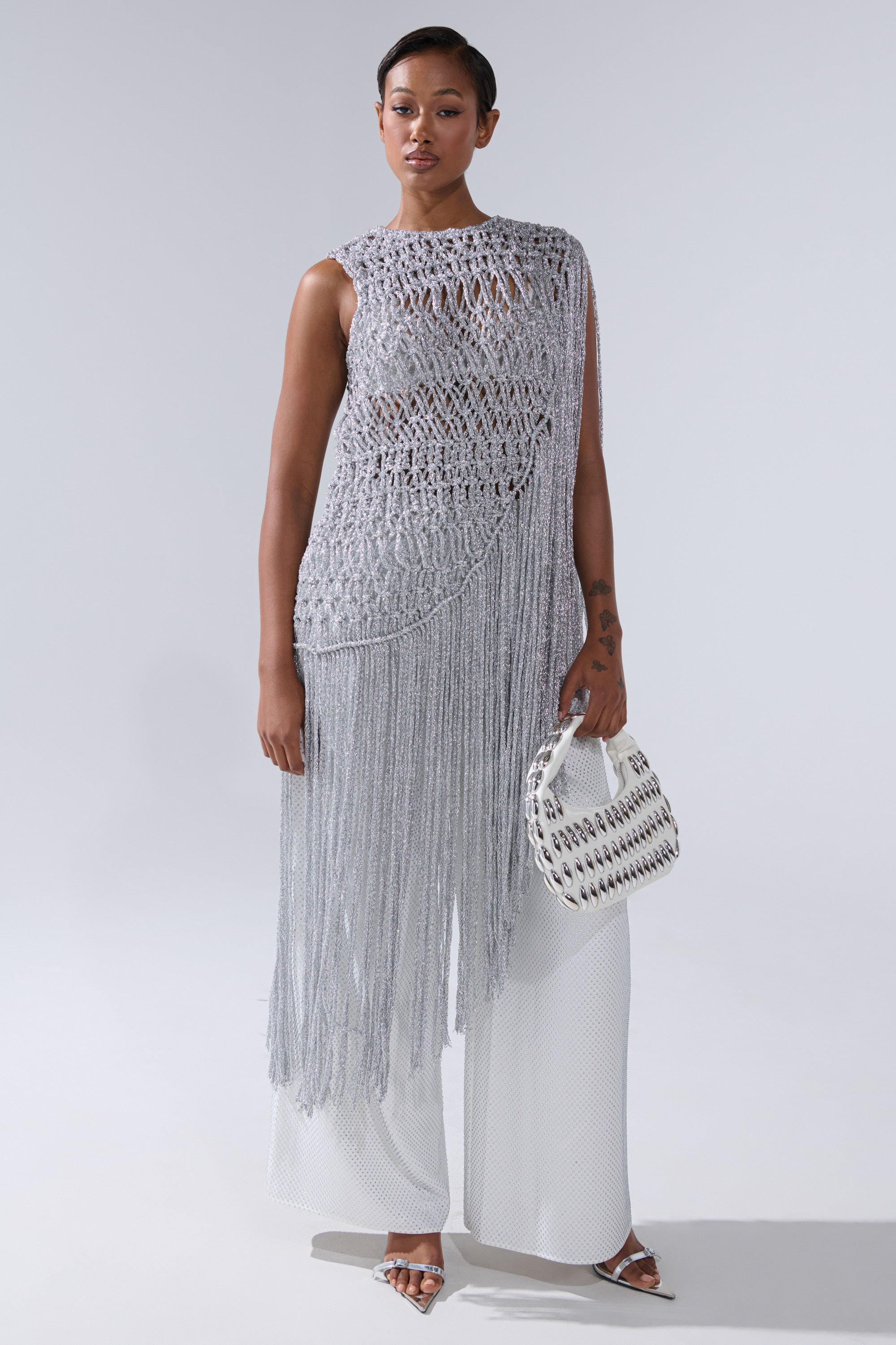Wearing the UNDRESS FRINGE BLOUSE—a silver, sleeveless, crocheted top with long fringe—paired with wide-leg white pants, silver heels, and a white woven handbag against a plain light background.