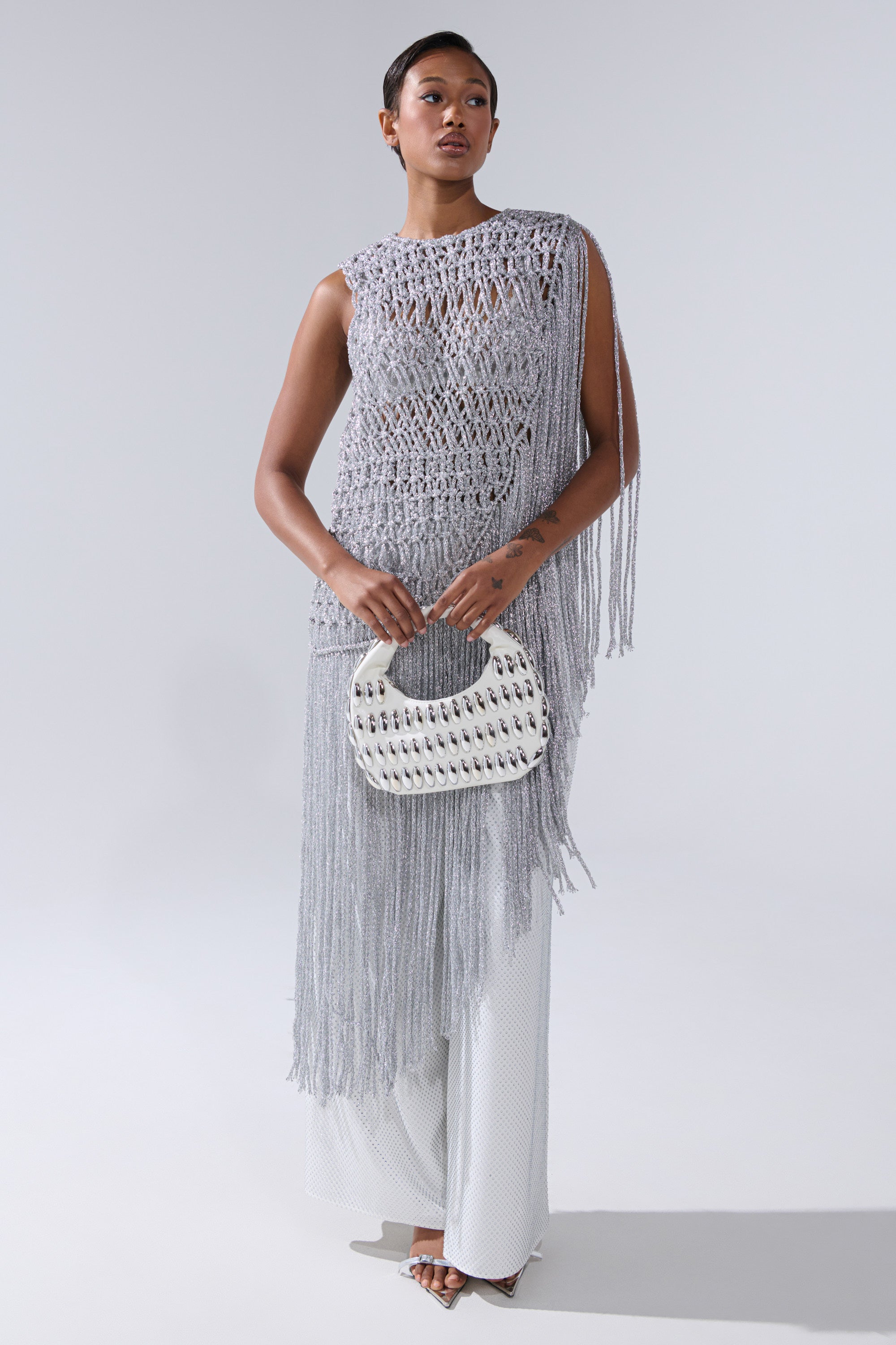 A woman poses in the UNDRESS FRINGE BLOUSE, a sleeveless gray crochet top with fringe, styled over white wide-leg pants and accessorized with a silver studded handbag and open-toe heels against a plain gray background.
