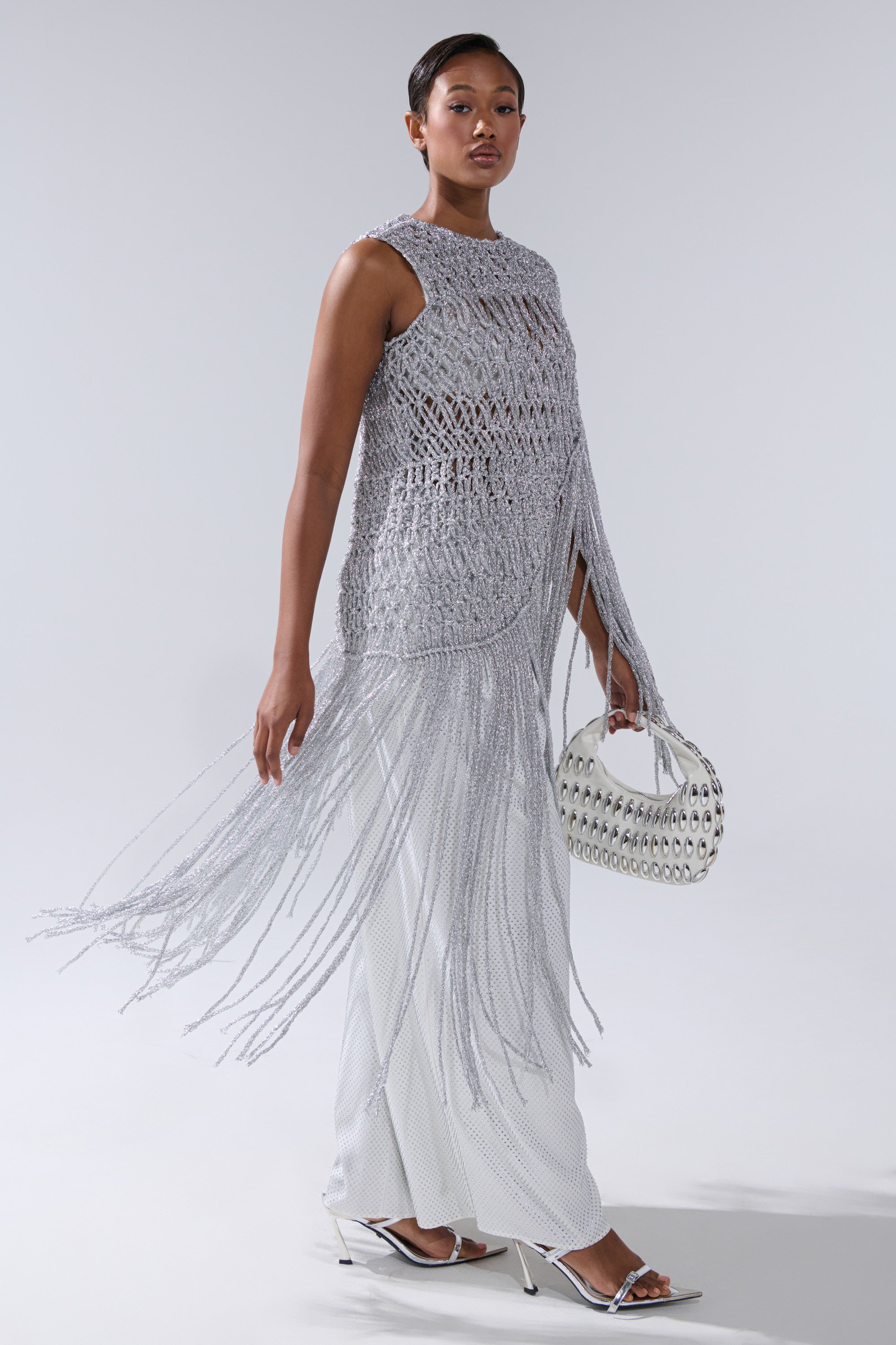A woman models the UNDRESS FRINGE BLOUSE, a sleeveless silver fringe top, styled over a white skirt with a white round handbag and heeled sandals, her hair slicked back against a plain light background.