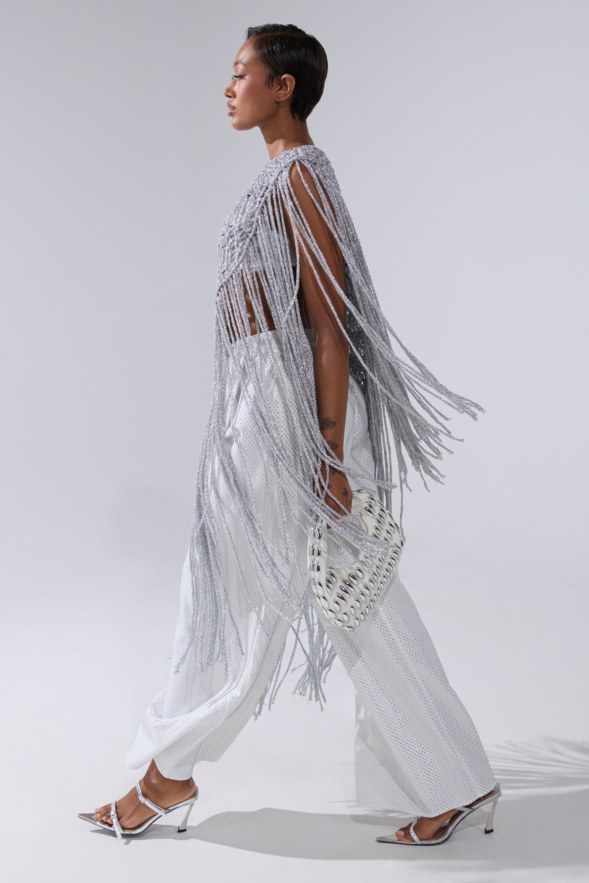 A person with short hair walks confidently in white heeled sandals, wide-leg textured pants, and the UNDRESS FRINGE BLOUSE, featuring dramatic long silver fringe. They carry a white woven handbag against a plain light background.