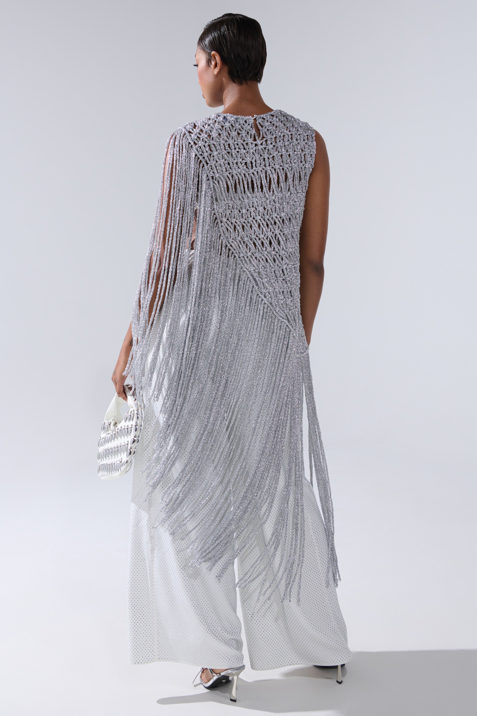 A person stands with their back to the camera in a long, silver UNDRESS FRINGE BLOUSE featuring crochet details and fringes, paired with white wide-legged pants and a small white handbag. The background is plain and light-colored.