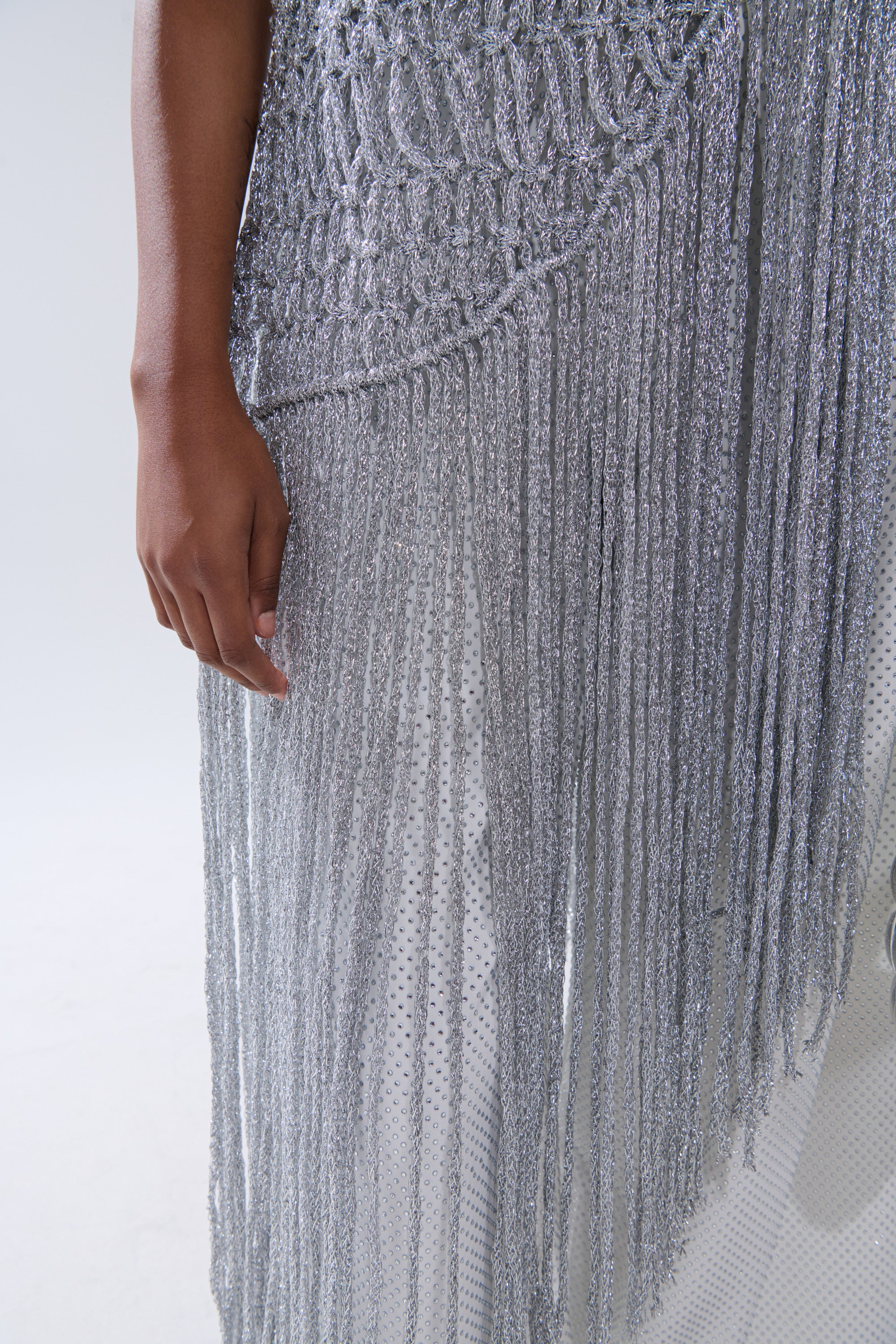 A person’s arm rests by their side, showcasing the UNDRESS FRINGE BLOUSE—textured silver with long, sparkling fringe and a mesh underlayer—set against a light background.