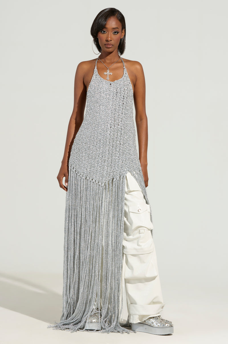 Wearing the ON SOME TOP, a silver fringed knit with an open-back design, they stand confidently alongside light cargo pants and shiny silver shoes. Their long dark hair complements a stylish necklace against a plain, light-colored background.