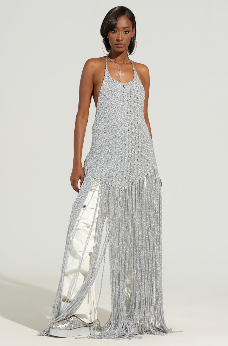 A person confidently stands against a plain background, wearing the ON SOME TOP, a silver, textured, sleeveless dress featuring an open-back design and floor-length fringe. The ensemble is perfectly complemented by shiny, metallic pants.
