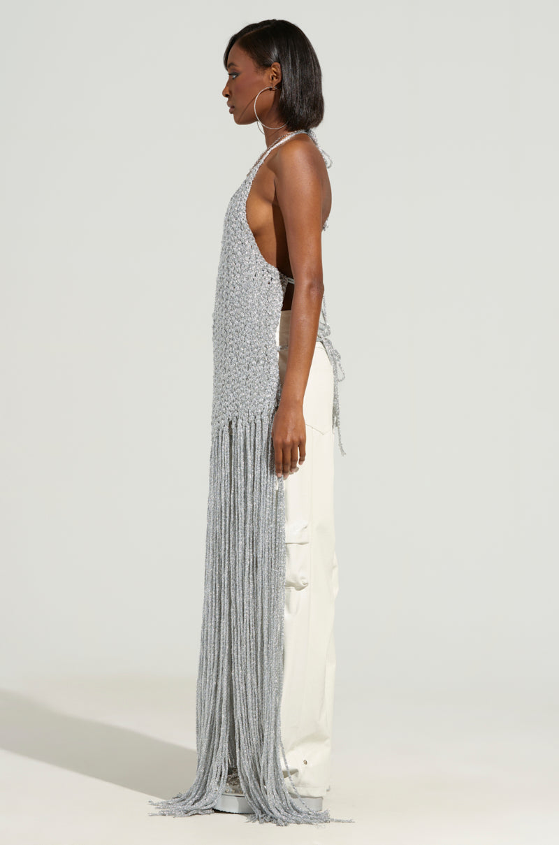 A person with shoulder-length hair stands in profile wearing the "ON SOME TOP," a long, light gray crochet top featuring an open-back design, paired with white pants. The fringe detail adds a charming touch, set against a plain, light-colored background.