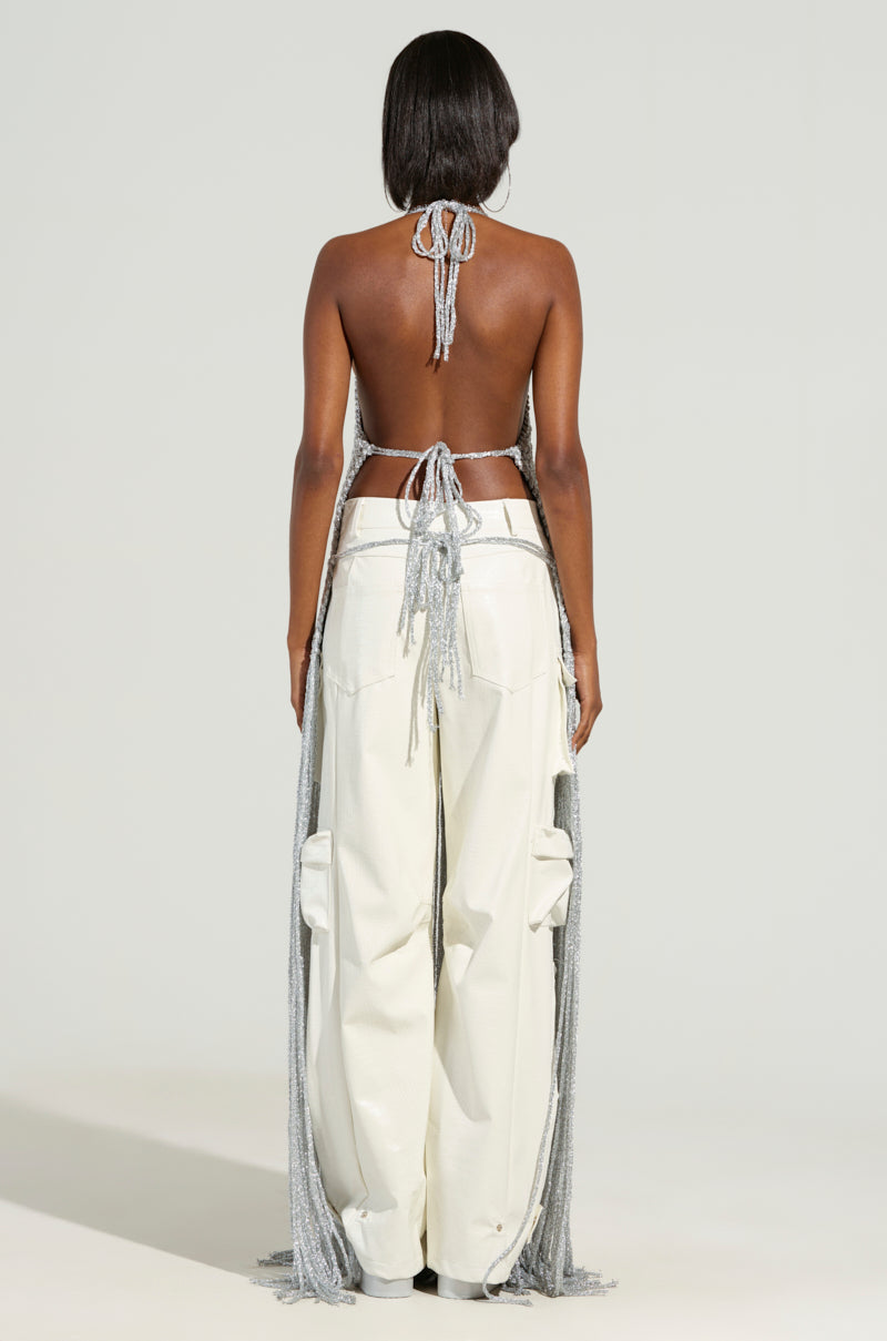 A person with long dark hair faces away from the camera, showcasing the ON SOME TOP—a halter-style design with an open back and floor-length fringe details—paired with cream-colored cargo pants. The setting is simple and light-toned.