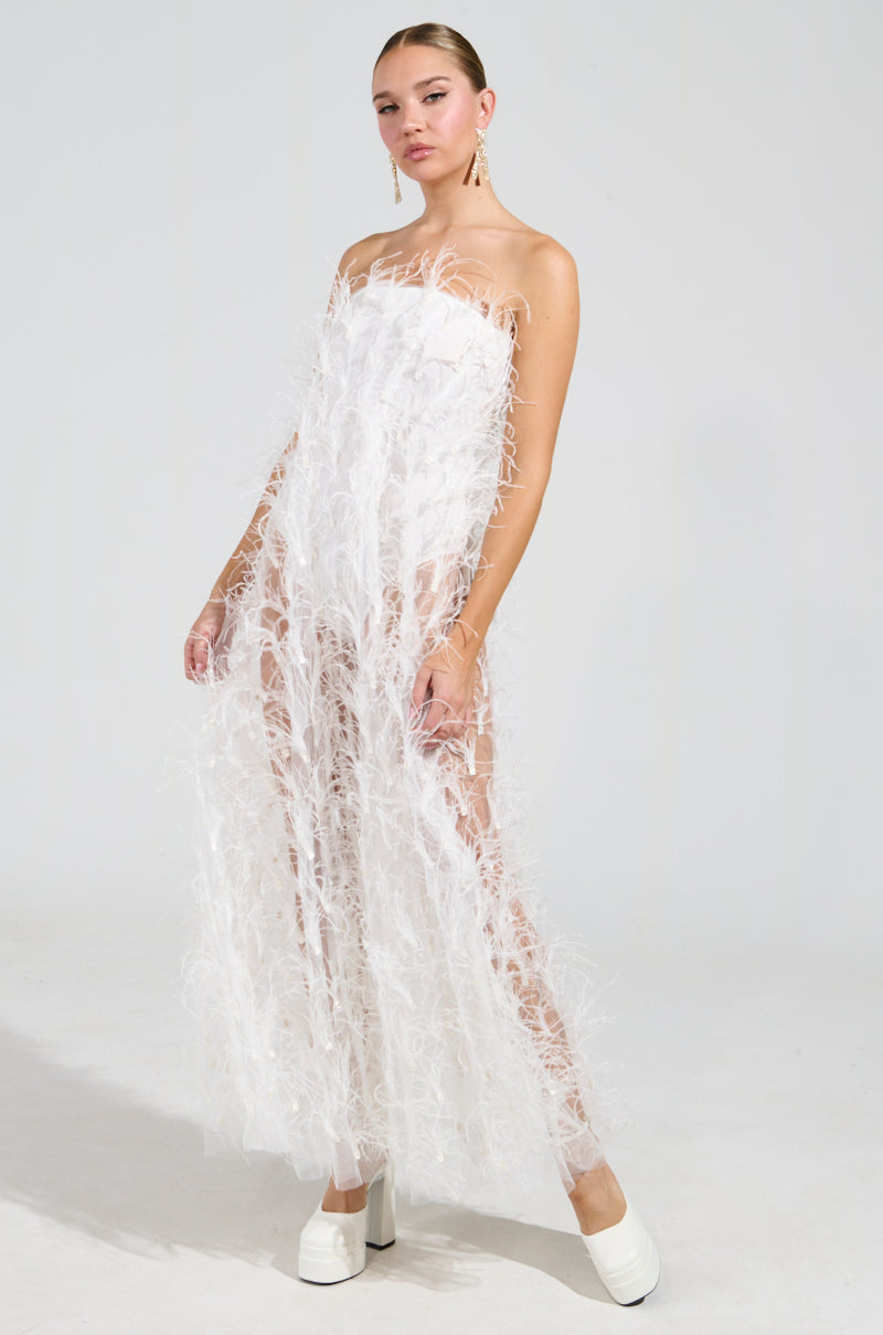 A woman stands confidently in the ONE OF A KIND FEATHER MAXI DRESS, featuring a strapless neckline and sheer tulle mesh with feather-like details. She pairs it with white platform shoes, has her hair pulled back, and wears long earrings against a plain, light-colored background.