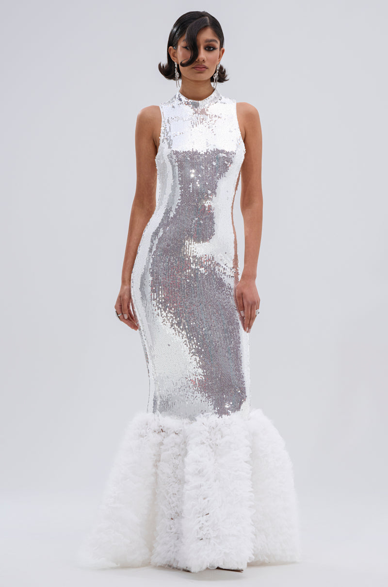 A woman stands against a plain light background wearing the BELLE OF THE BALL SEQUIN RUFFLE MAXI DRESS IN WHITE, a sleeveless, form-fitting gown with a high neckline and dramatic ruffled hem that highlights its mermaid silhouette.