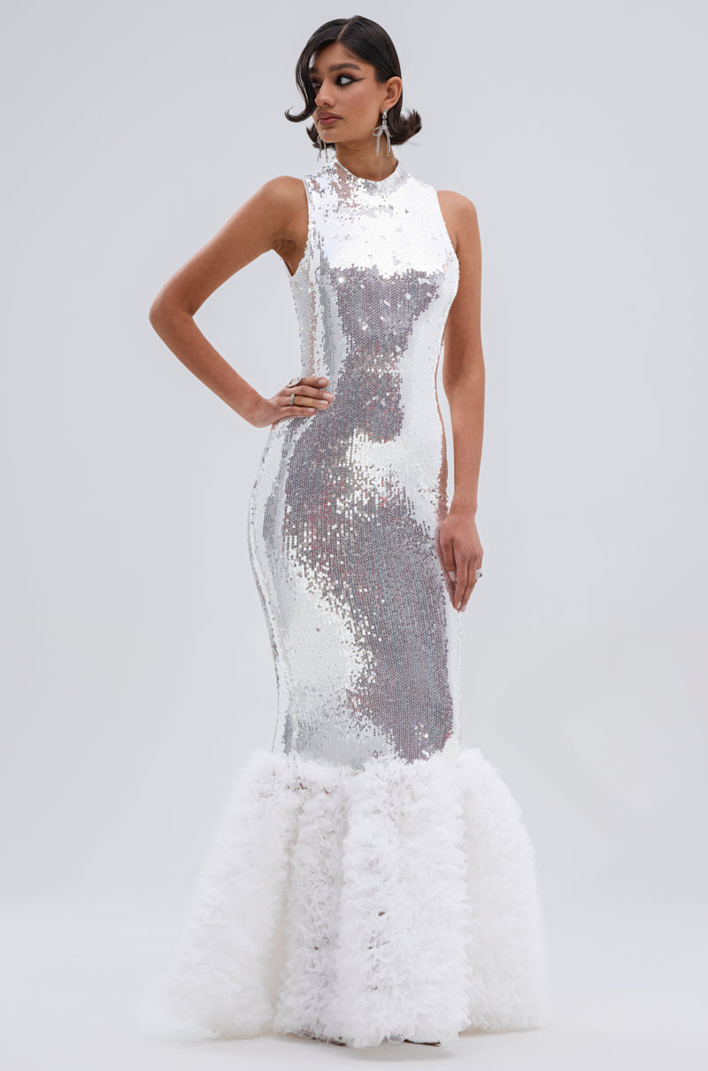 A woman poses in the BELLE OF THE BALL SEQUIN RUFFLE MAXI DRESS IN WHITE, a sleeveless maxi with a mermaid silhouette, tonal sequins, high neckline, and dramatic ruffled hem, against a plain light gray background.
