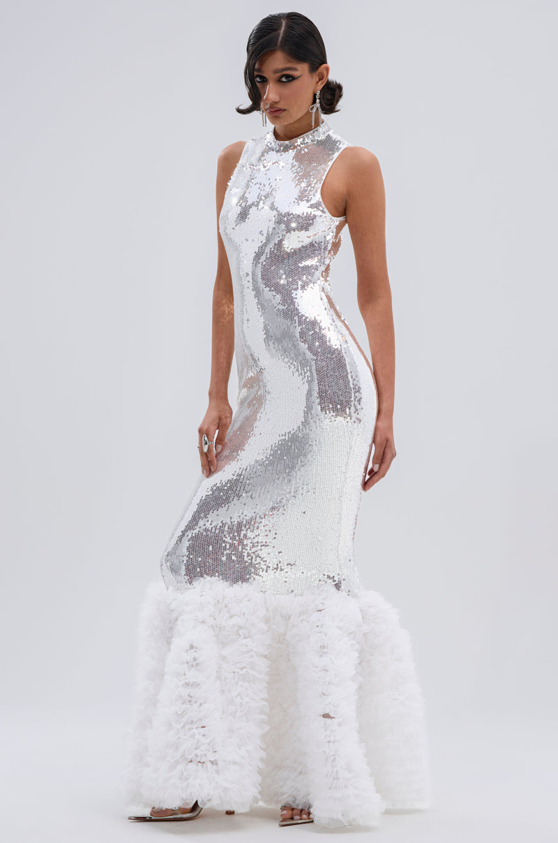 A woman with short dark hair poses against a plain light background wearing the BELLE OF THE BALL SEQUIN RUFFLE MAXI DRESS IN WHITE, featuring a sleeveless design, high neckline, sequins, and a dramatic voluminous ruffled hem.