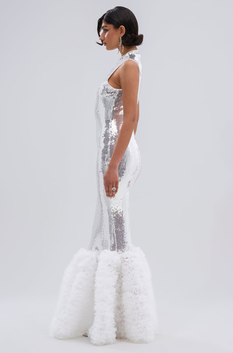 A woman stands in profile against a plain light gray background, wearing the BELLE OF THE BALL SEQUIN RUFFLE MAXI DRESS IN WHITE, featuring a sleeveless fitted mermaid silhouette with dramatic sequin and ruffle details at the hem.