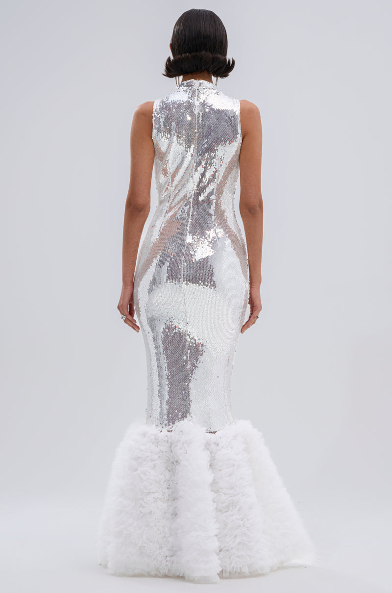 A person with short, dark hair is seen from behind, wearing the BELLE OF THE BALL SEQUIN RUFFLE MAXI DRESS IN WHITE. The mermaid silhouette features tonal sequins and a fluffy ruffled hem, shimmering against a plain light gray background.