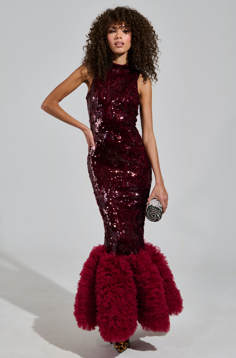 A woman poses in a sleeveless, burgundy BELLE OF THE BALL SEQUIN RUFFLE MAXI DRESS, featuring a voluminous ruffled hem that evokes a Mermaid Silhouette. She accessorizes with a small, round clutch and leopard print shoes. Her curly hair frames her face as she stands against a gray background.