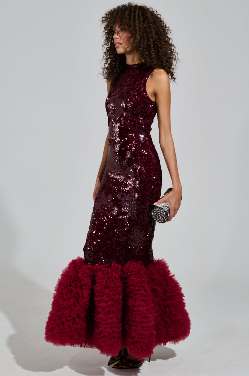 A woman in a sleeveless, dark red BELLE OF THE BALL SEQUIN RUFFLE MAXI DRESS with a ruffled hem stands against a plain background. She holds a small, patterned clutch and has curly hair, exuding elegance.
